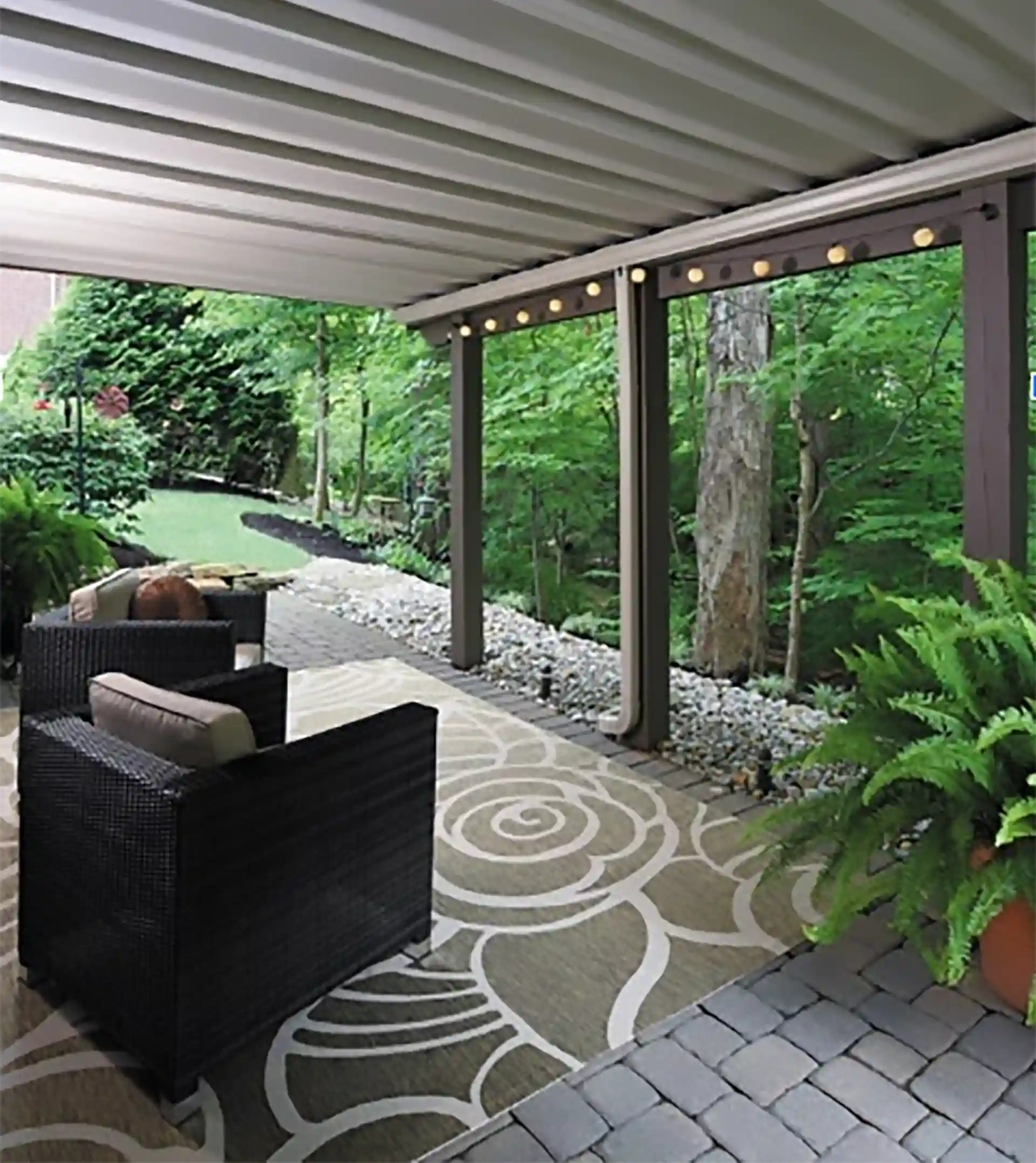 Underdeck view of a covered patio with modern furniture, pavers, and hanging lights, surrounded by greenery and trees.