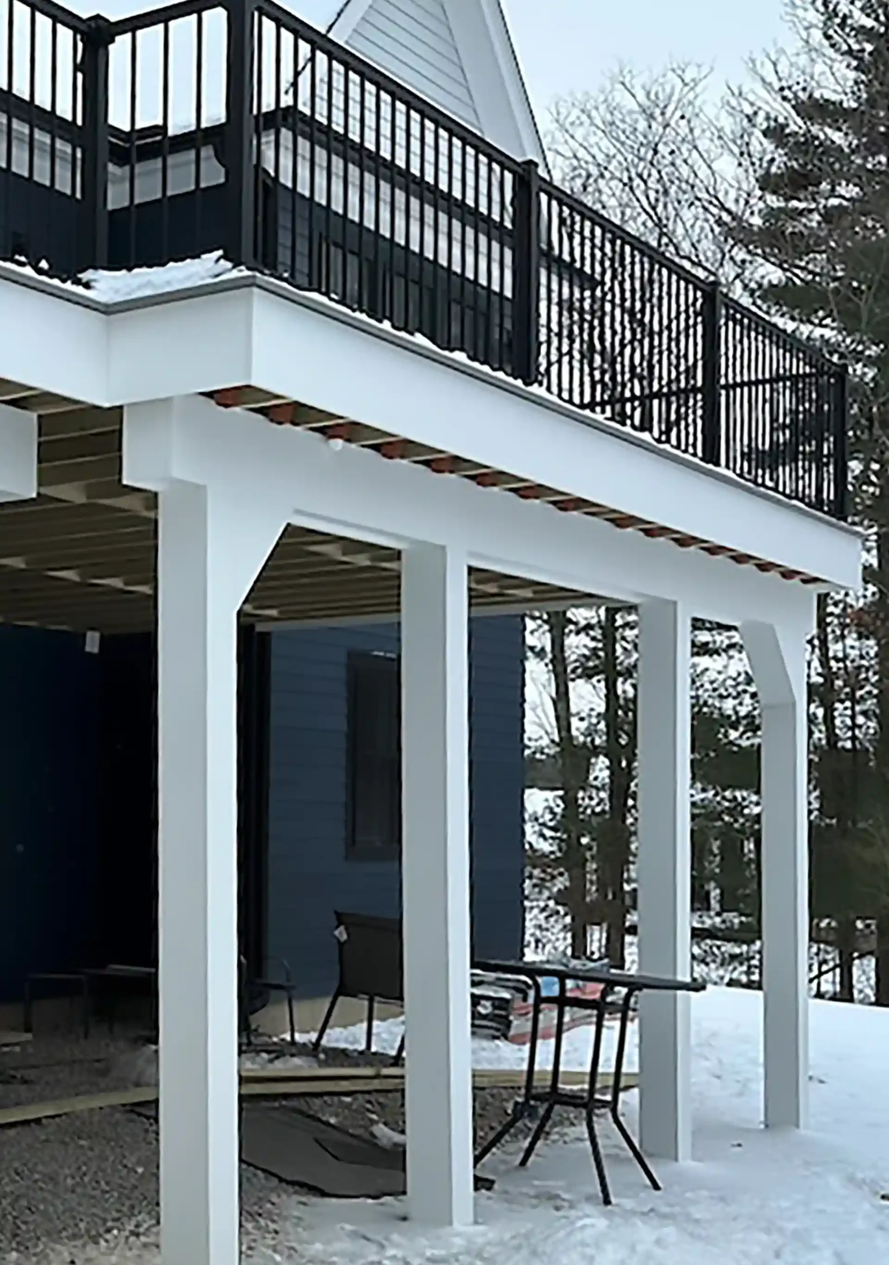 Under-deck view showing a waterproofing system installed beneath a raised deck with a black railing and snow-covered ground.