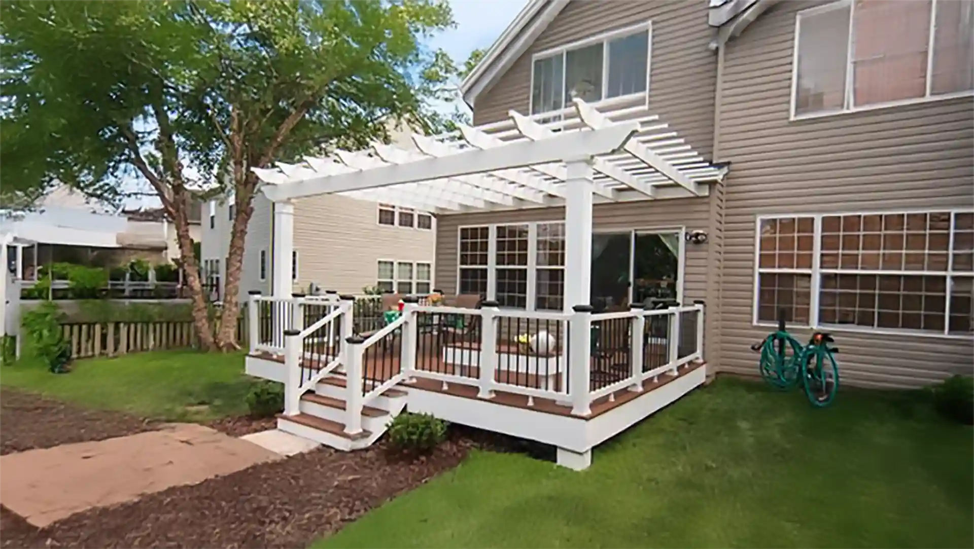 Backyard deck with white pergola, railing with black balusters, steps leading to lawn, and patio furniture. 