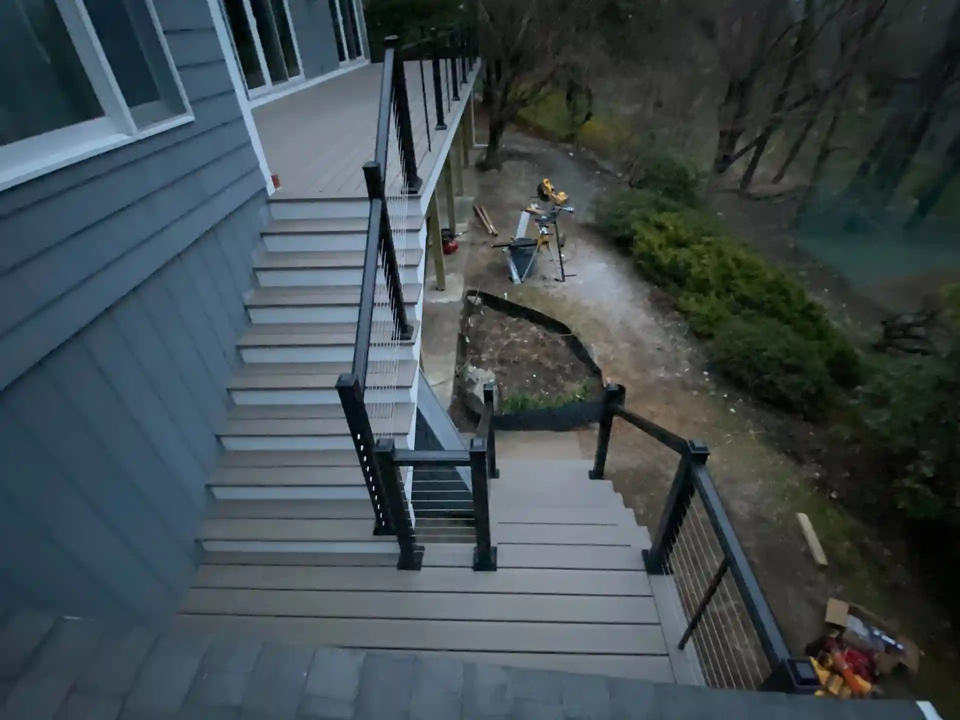 A multi-level deck with cable railings and stairs leading from the upper deck to the lower level, with visible landscaping and construction tools.
