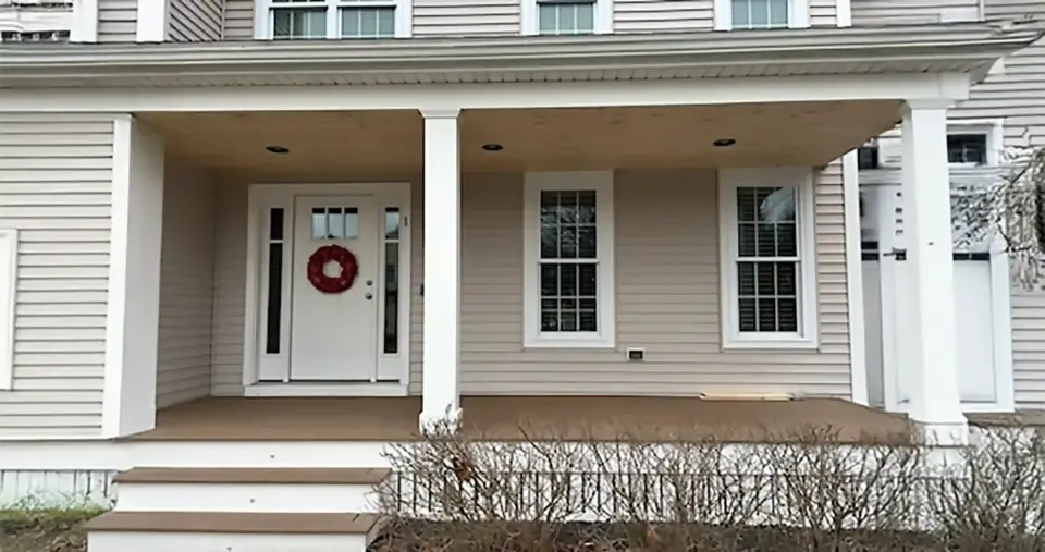 Front porch with white door, two windows with shutters, two white columns supporting roof, and wooden deck floor.